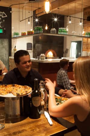 Two people sharing a pizza and bottle of wine, holding hands and looking at each other across the table.