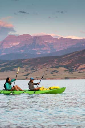 Two women kayaking on a lake, with mountains illuminated pink by the setting sun in the background.